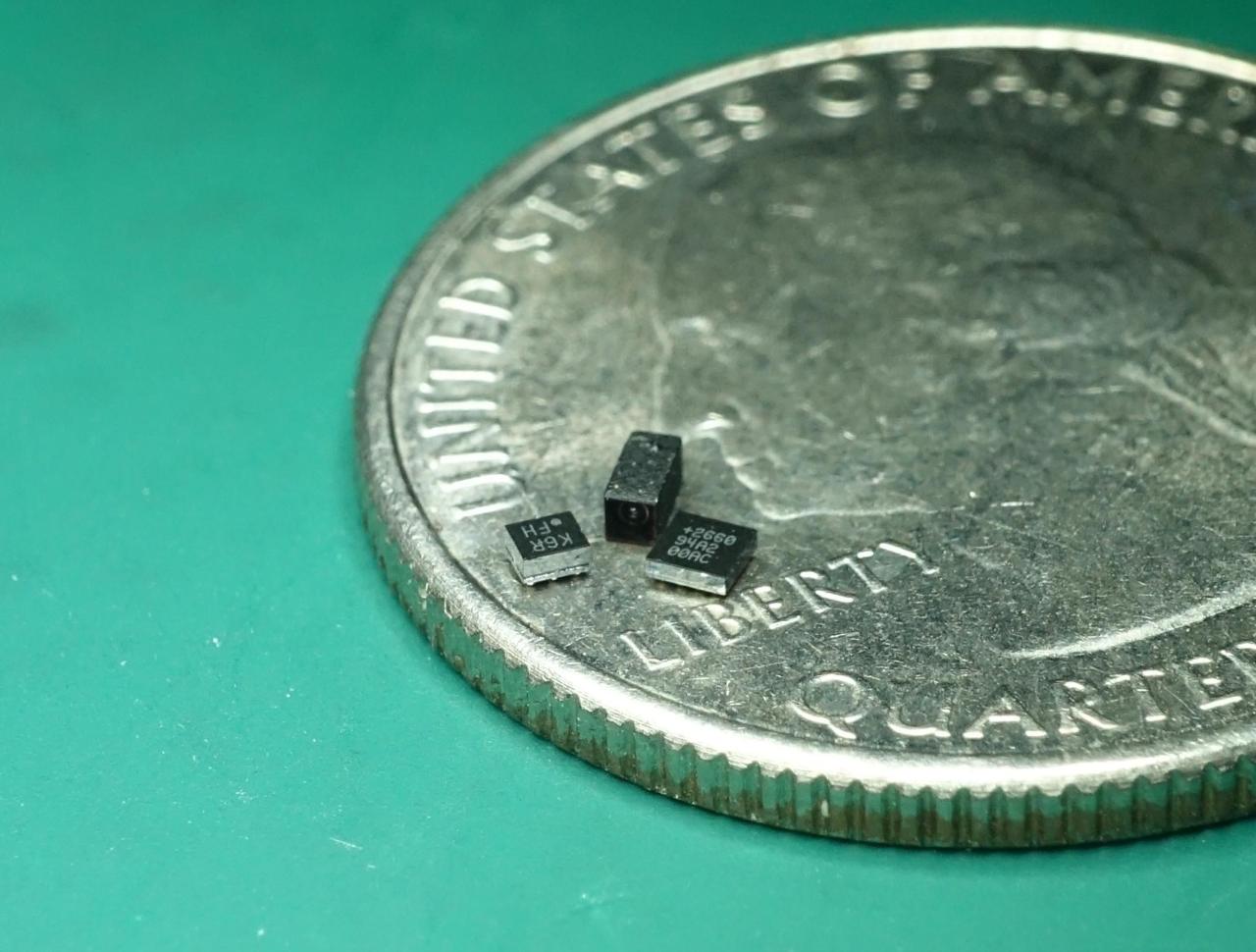 A photo of three tiny black electronic components on a US quarter, where each component is approximately the size of a letter on the coin